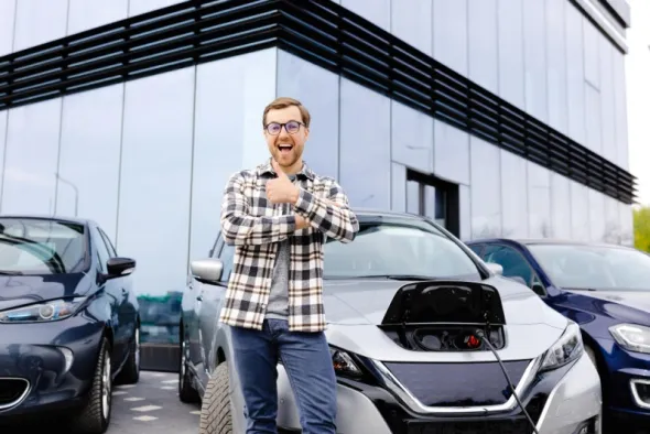 Zadowolony mężczyzna w koszuli w kratę stojąc na parkingu wypożyczalni samochodów podnosi w górę kciuk
