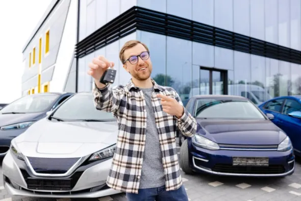 Mężczyzna w koszuli w kratę stojąc na parkingu dumnie pokazuje kluczyki od wynajętego samochodu 