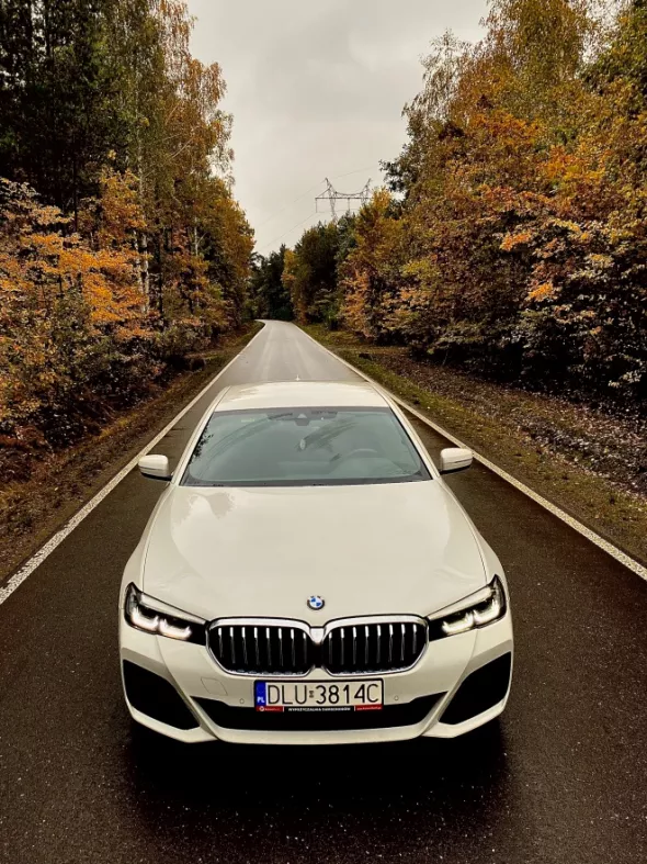 Białe BMW 5 to na tle drogi wśród jesiennych drzew to samochód premium chętnie wynajmowany w Kaizen Rent 