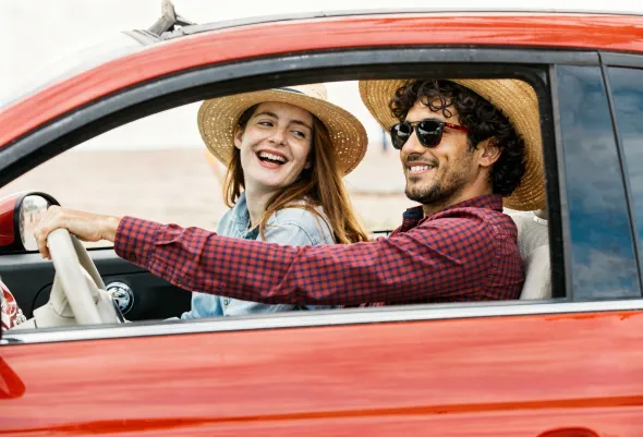 Młoda para jedzie czerwonym czerwonym samochodem. Kobieta uśmiechem poprawia kapelusz, mężczyzna prowadzi radośnie wynajęte auto