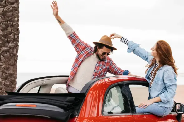 Radosna para wychylająca się z okien jadącego czerwonego auta, machająca rękami na pożegnanie; dynamiczne ujęcie pełne energii i entuzjazmu.