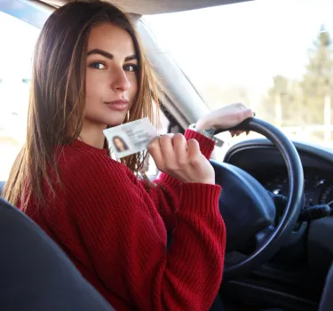 Młoda kobieta w czerwonym swetrze za kierownicą samochodu pokazuje swoje prawo jazdy